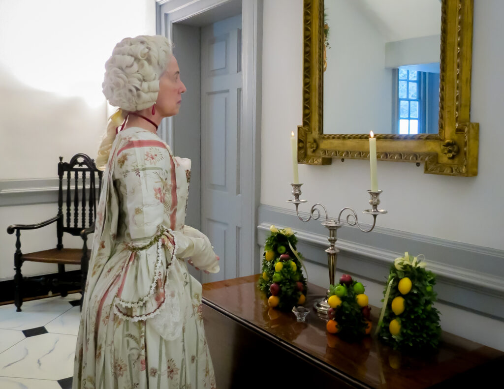 In the Central Passage. A reenactor portraying a wealthy colonial woman stands before a mirror in the Paca House. 2025.