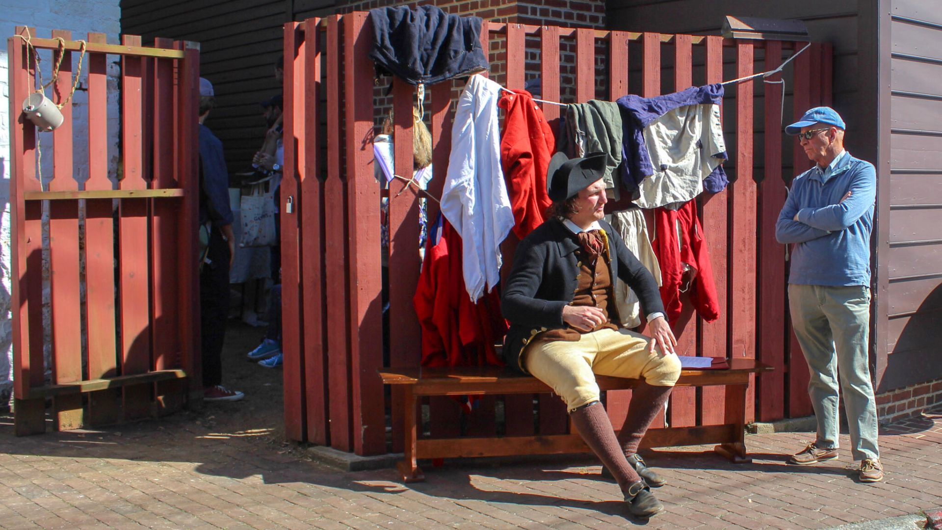 A reenactor explains the 18th-century secondhand clothing trade to a visitor in a living history photography image.
