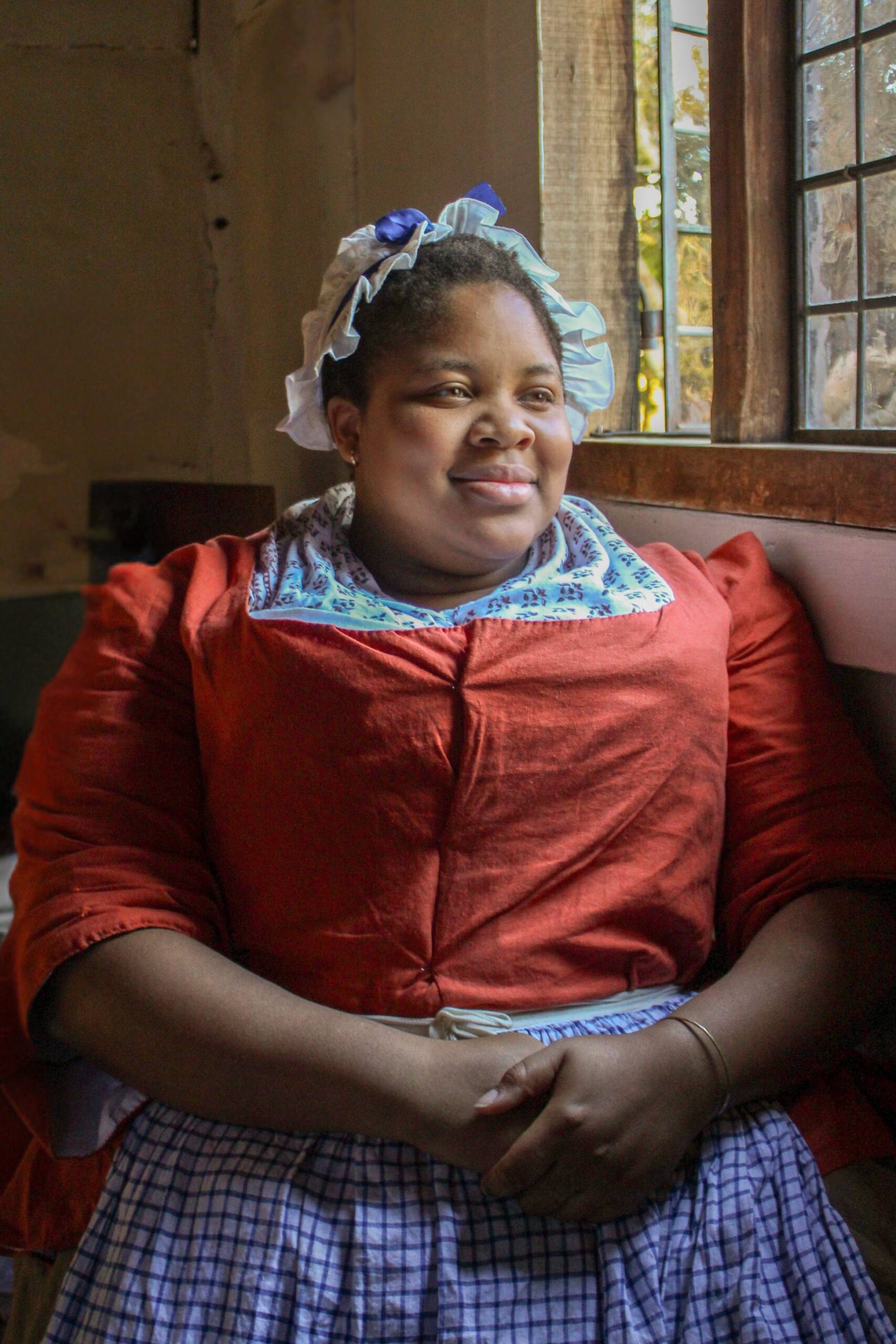 Living history photography portrait of a Black woman reenactor seated in front of a window, wearing 18th-century clothing.