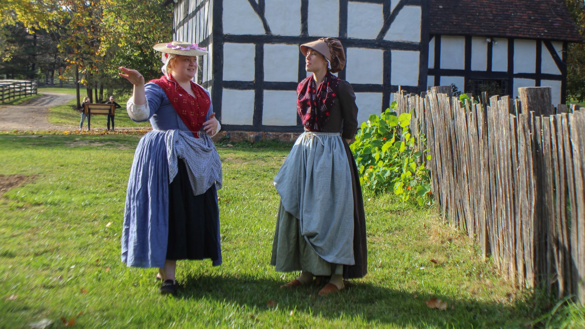 Living history photography shows two 18th-century camp follower reenactors outside a Tudor-style structure.